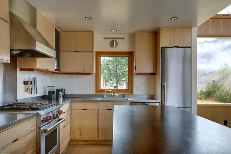 The kitchen features wood cabinetry, stainless steel countertops, and a framed window overlooking a natural landscape.
