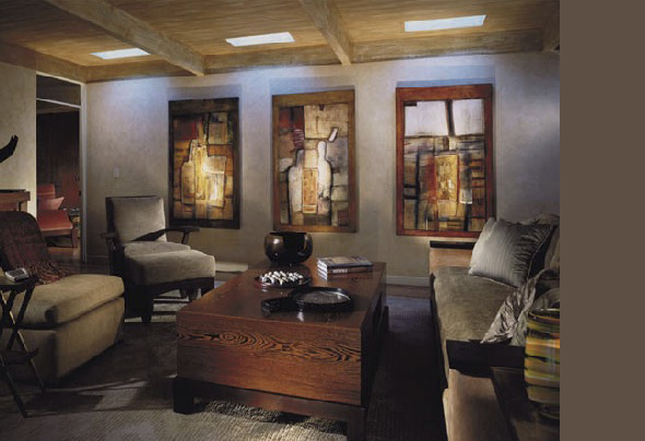 A dimly lit living room with a wooden coffee table, upholstered chairs, and three abstract paintings on the wall under exposed ceiling beams