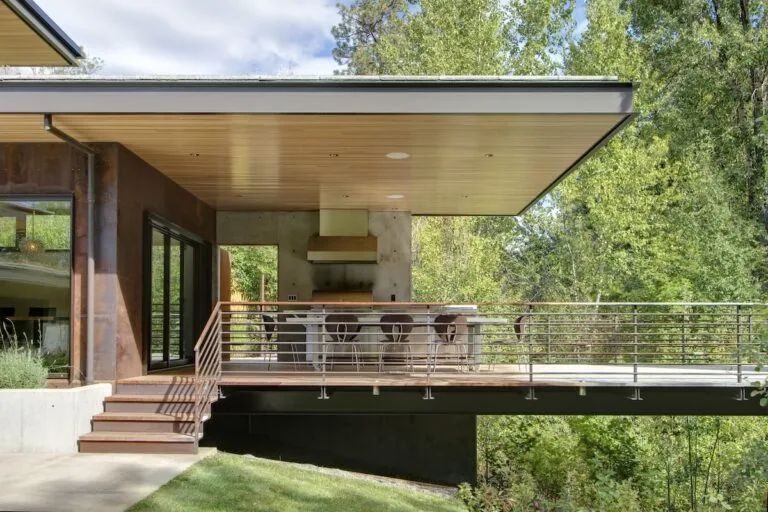 Covered outdoor patio with wood-paneled ceiling and metal stair railing at Montana Creekside Residence.