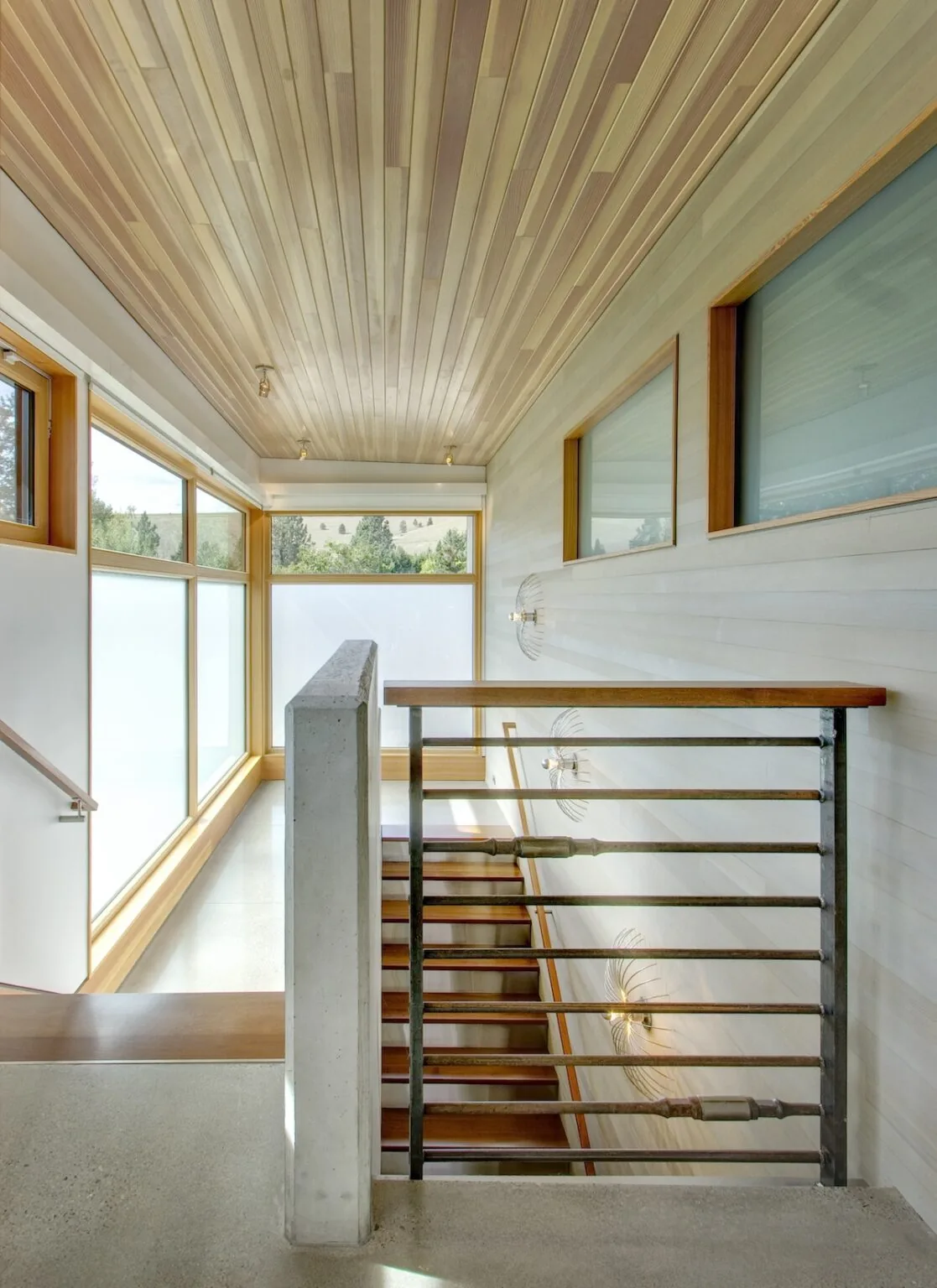 Stairwell with wood ceiling and metal railing illuminated by wall sconces, featuring large windows with frosted glass