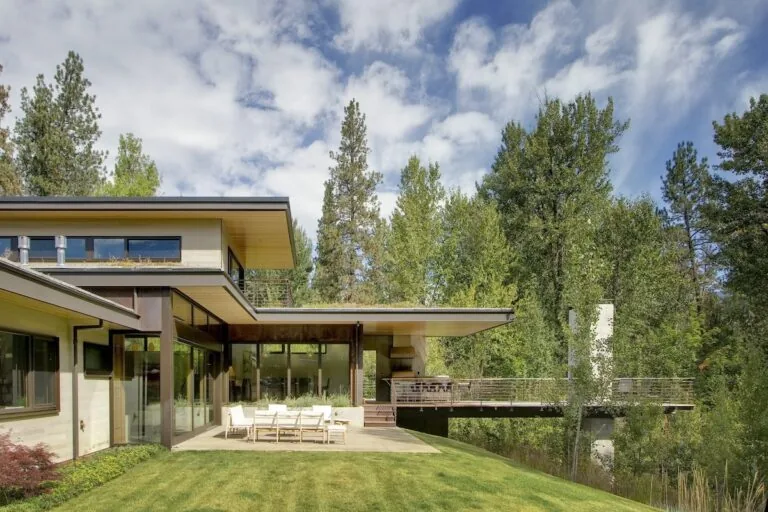 Montana Creekside Residence features a flat roof, floor-to-ceiling glazing, and a cantilevered deck above a creek.