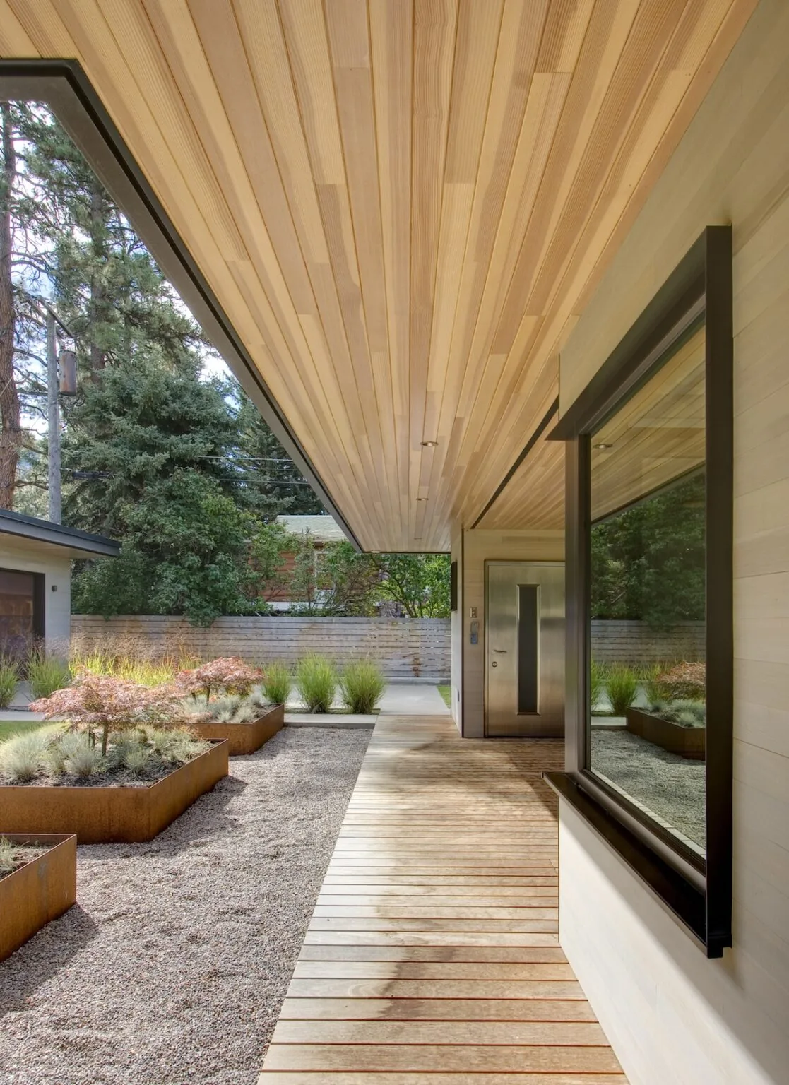 The Montana Creekside Residence features a wood-paneled ceiling over the exterior walkway leading to a modern entry door.