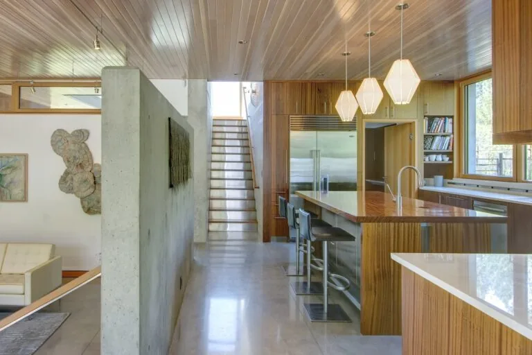 Montana Creekside Residence kitchen features wood cabinetry, a central island with wood top, and a stairway behind a concrete wall.