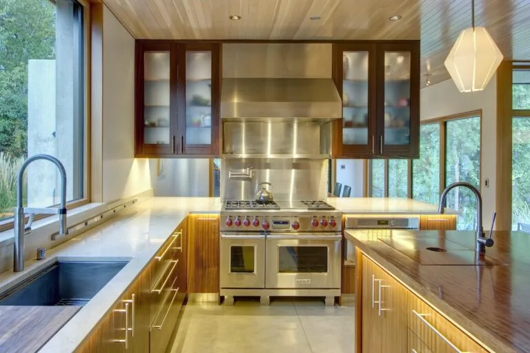 Modern kitchen with wood-paneled ceiling, stainless steel stove and hood, and large windows overlooking greenery.