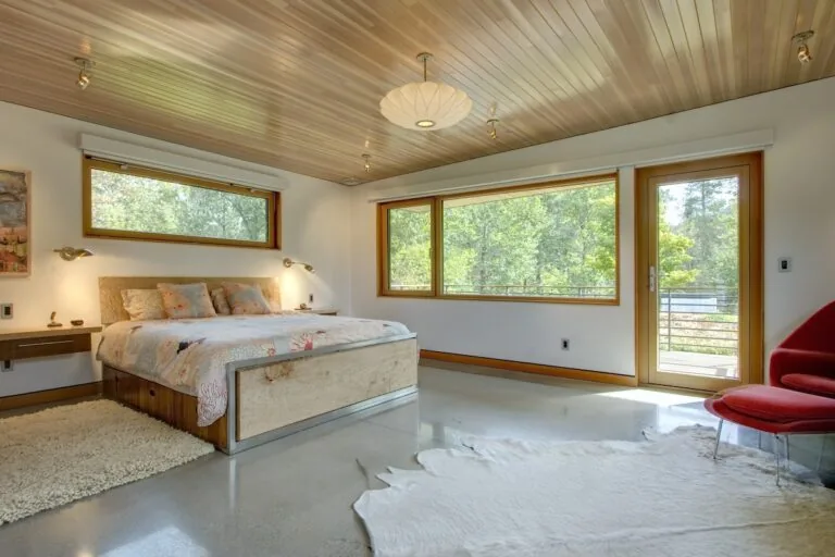 Montana Creekside Residence bedroom features wood ceiling, large wood-framed windows, and polished floor with area rugs.