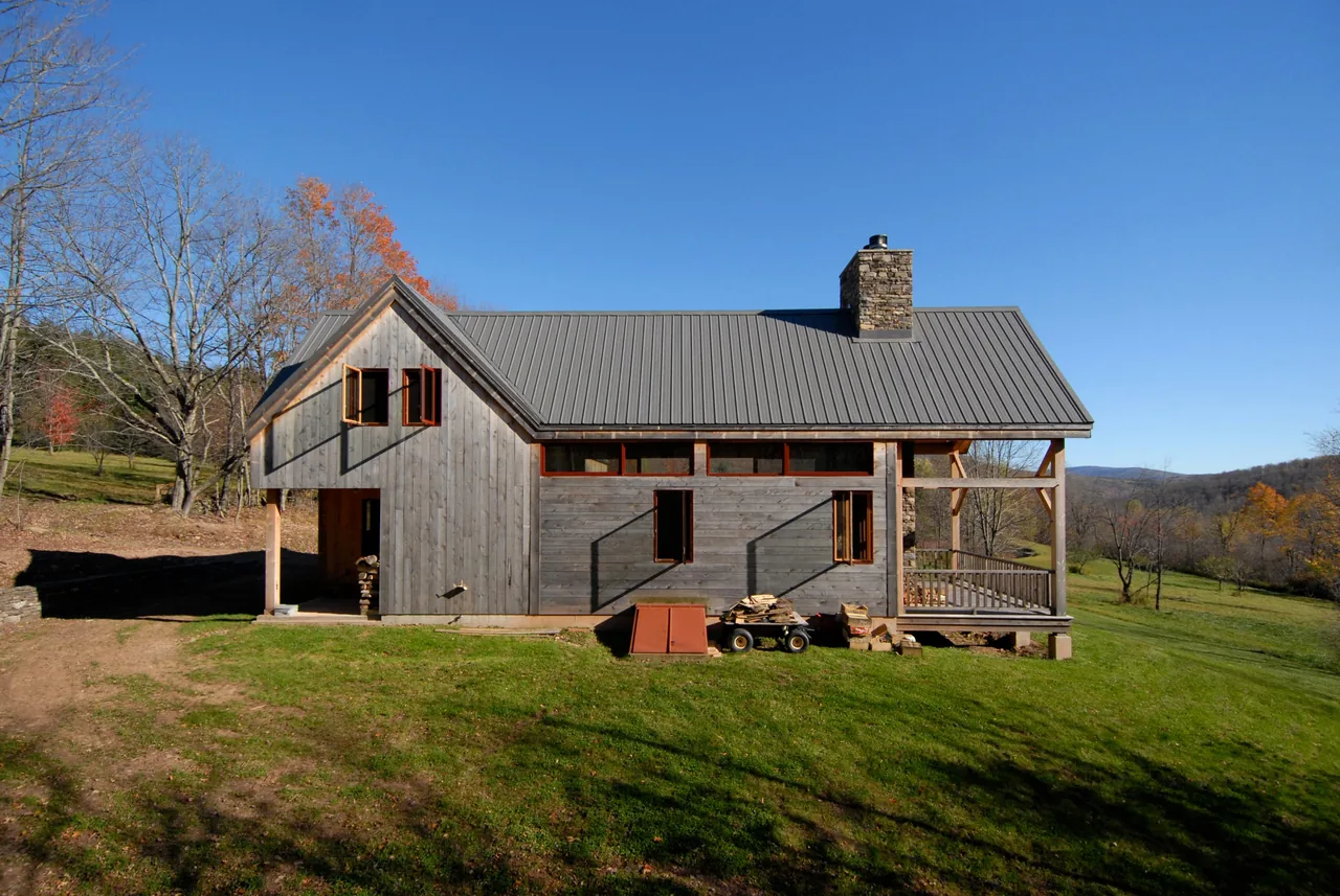 The Catskills Retreat features wood siding, a standing seam metal roof, and a stone chimney in a rural landscape.