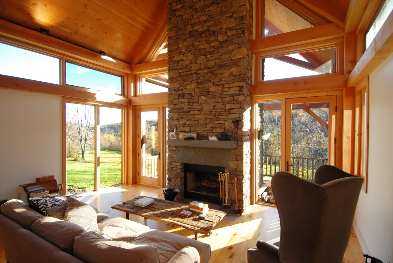 The Catskills Retreat living room features a stone fireplace with wood trim and expansive windows revealing timber porch details.