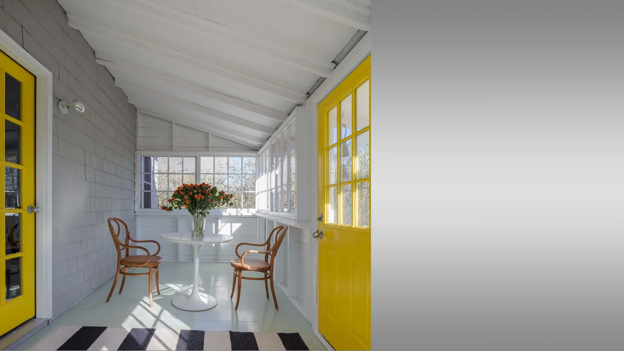 Enclosed porch space with white walls, yellow door, wooden chairs, and floral centerpiece on table.