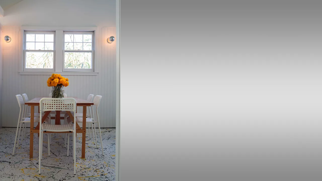 Dining area at Spagnola House with a wooden table, white chairs, and orange flowers under two windows.
