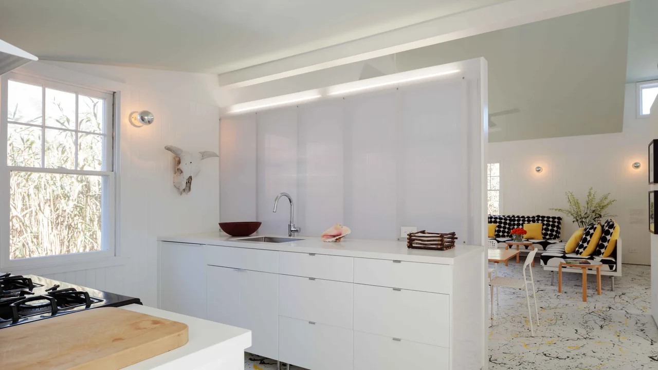 Interior view of the Spagnola House kitchen with white cabinetry and a sink counter, opening to a living space featuring patterned seating and yellow pillows.