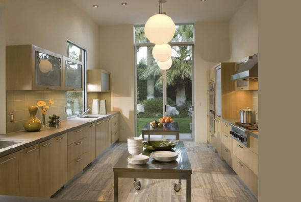 Modern kitchen featuring light wood cabinetry, a stainless steel central table, and pendant globe lighting with garden views.
