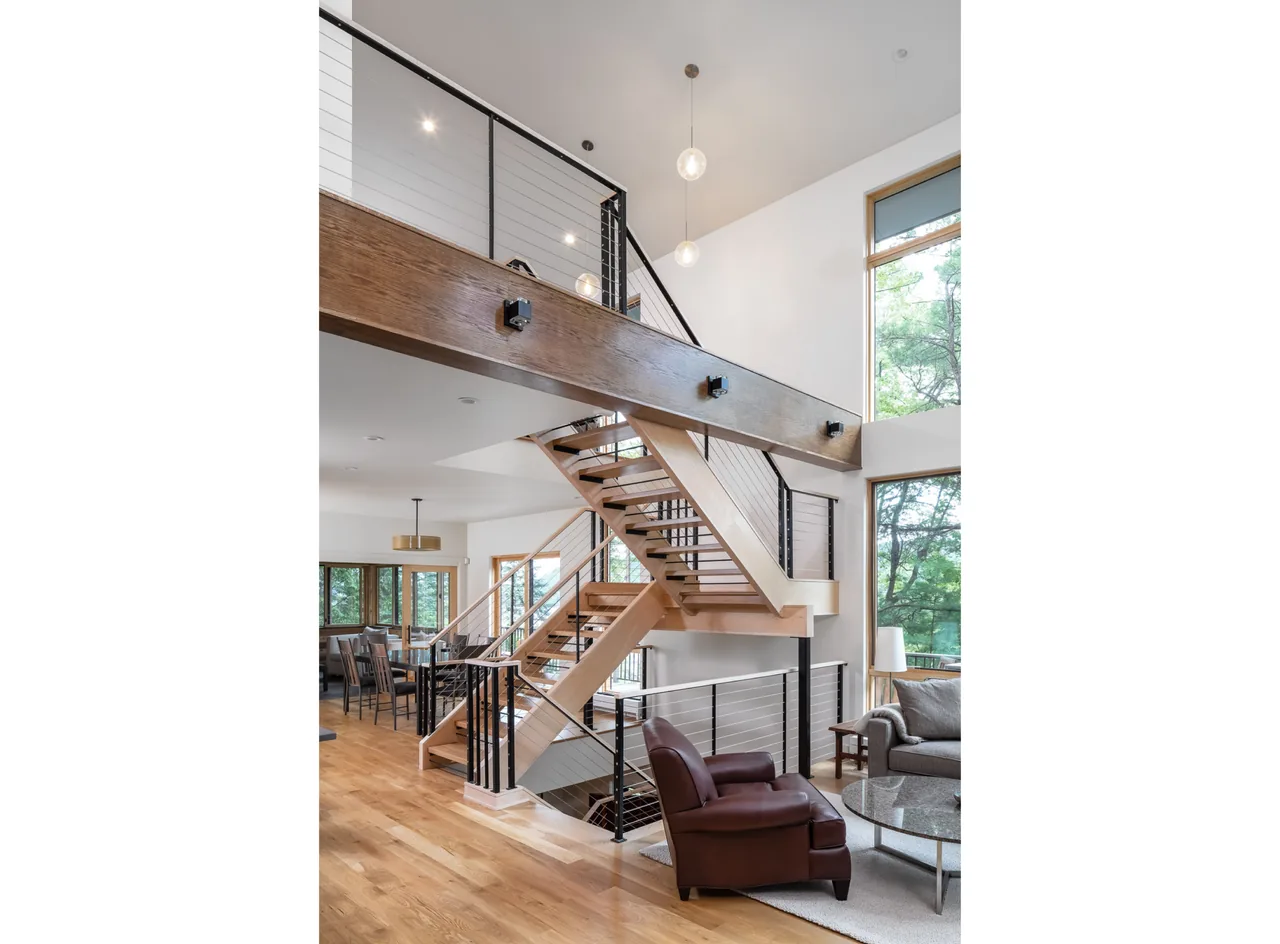 Modern living area with a central wood and cable railing staircase, floor-to-ceiling windows, and simple pendant lighting.