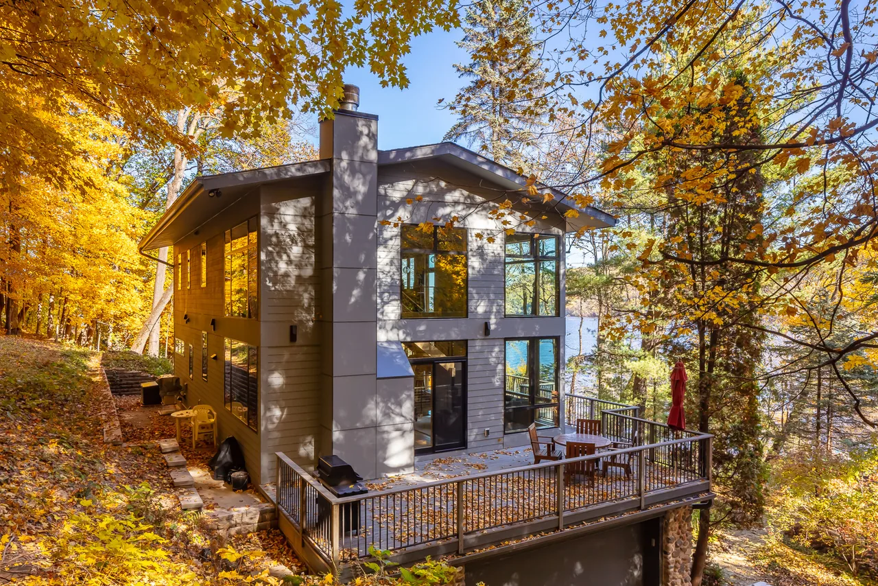 River Residence features floor-to-ceiling windows and a large outdoor deck overlooking the river, set in a wooded area in fall.