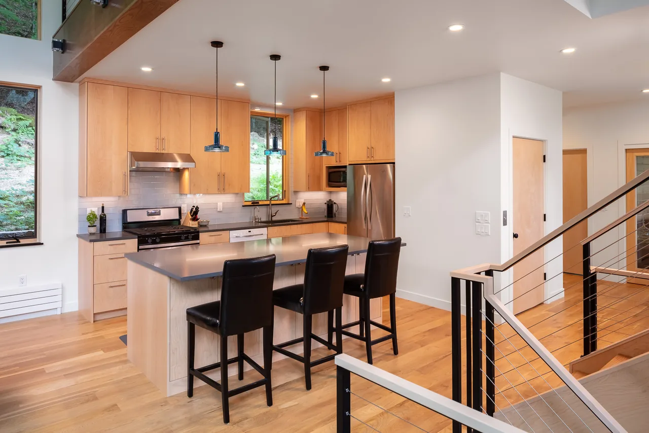 The River Residence kitchen features light wood cabinetry, a central island with seating, and stainless steel appliances.