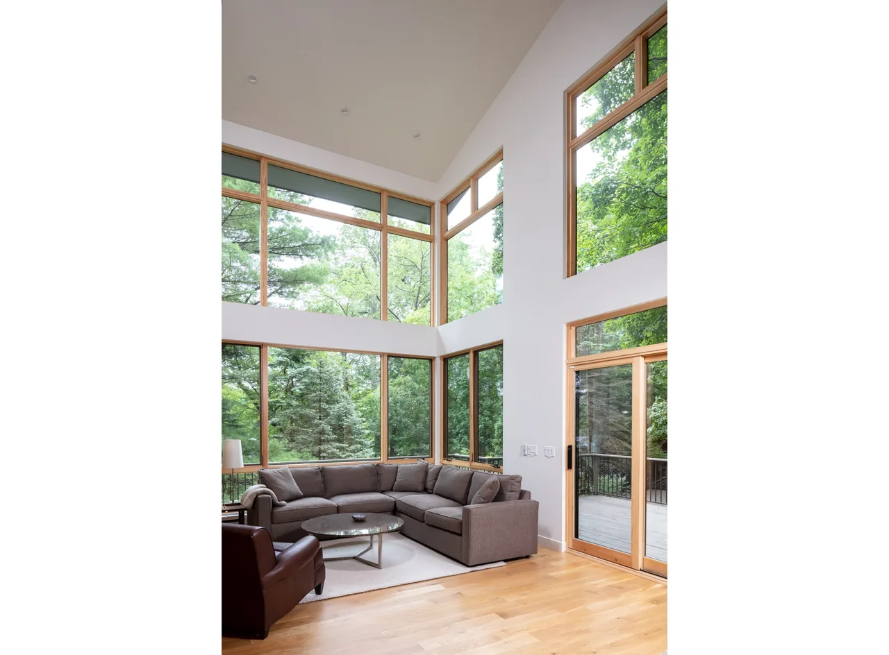 Tall-ceiling living room with floor-to-ceiling windows overlooking lush trees and a deck outside.