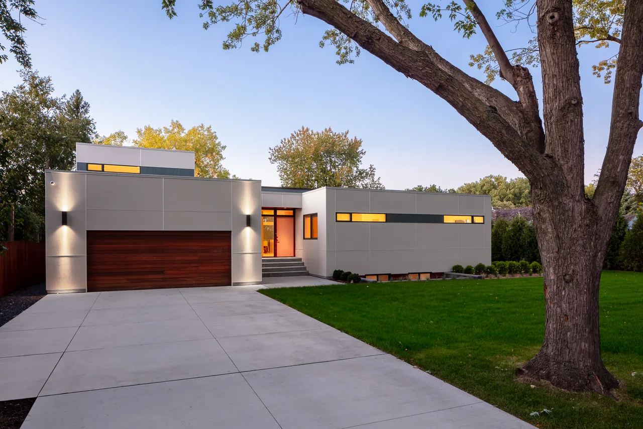 Modern two-story house with gray panel siding, wood garage door, and horizontal windows featuring exterior lighting at dusk.