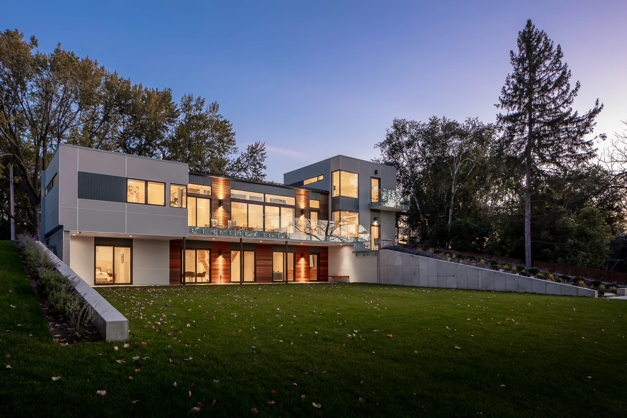 Modern two-story house with large glass windows overlooking a green lawn and surrounded by mature trees at dusk.