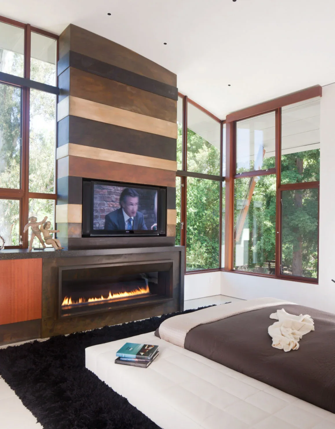 Bedroom features a built-in linear fireplace under a wall-mounted TV in front of large wood-framed corner windows showing trees outside.