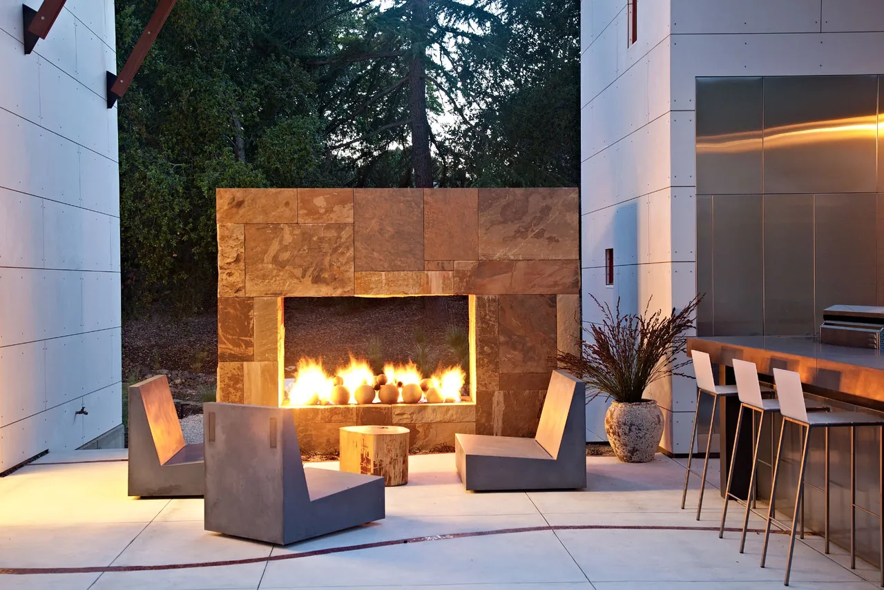 The Saratoga Creek House outdoor living area features a stone fireplace with concrete chairs and a bar counter with stools.