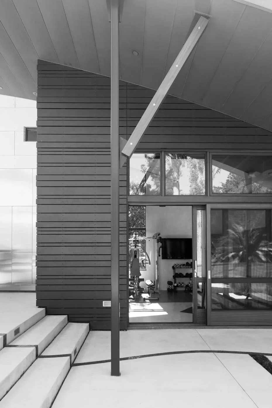 The Saratoga Creek House features a curved metal roof, wide exterior concrete stairs, and glass sliding doors leading to a home gym.