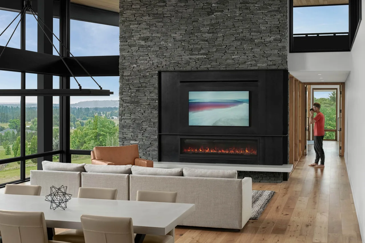 Modern living room with a gray stacked stone fireplace wall, surrounded by floor-to-ceiling windows and an open view to the landscape outside.