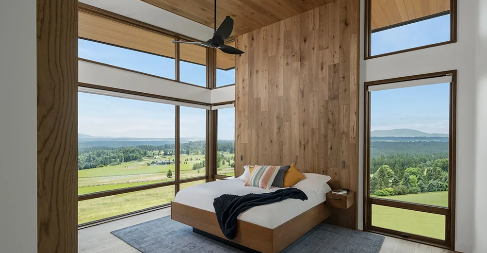 Bedroom with floor-to-ceiling glazing offers panoramic views of a green valley and distant hills.