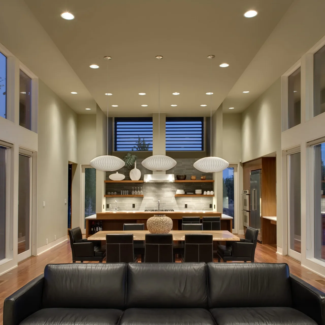 The Fadjur Lane Residence features a high-ceiling living and dining space with black leather seating and an open kitchen with wood cabinetry.