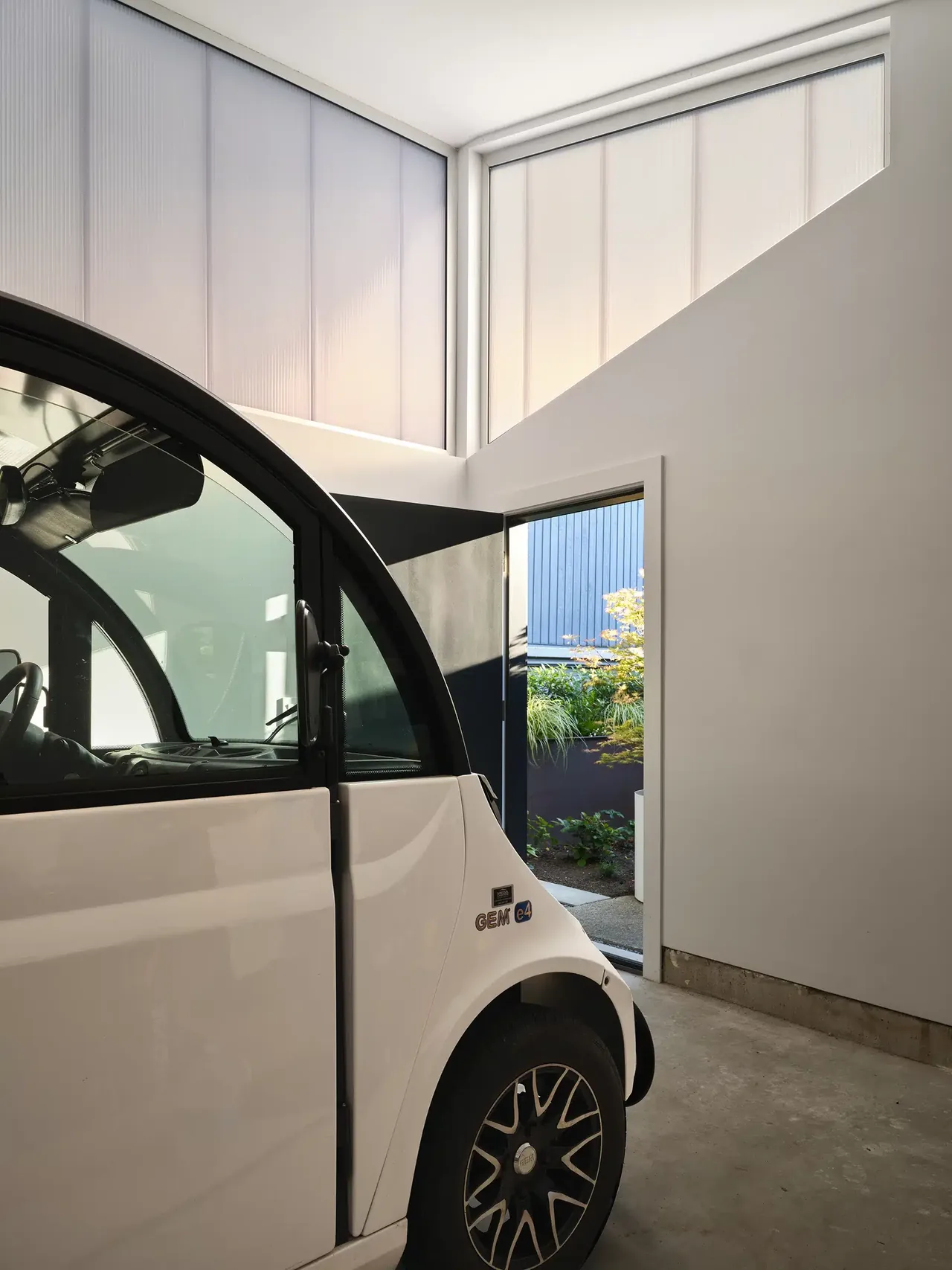 Garage interior showing a small electric car parked near an open door leading to a garden with plants and outdoor light.