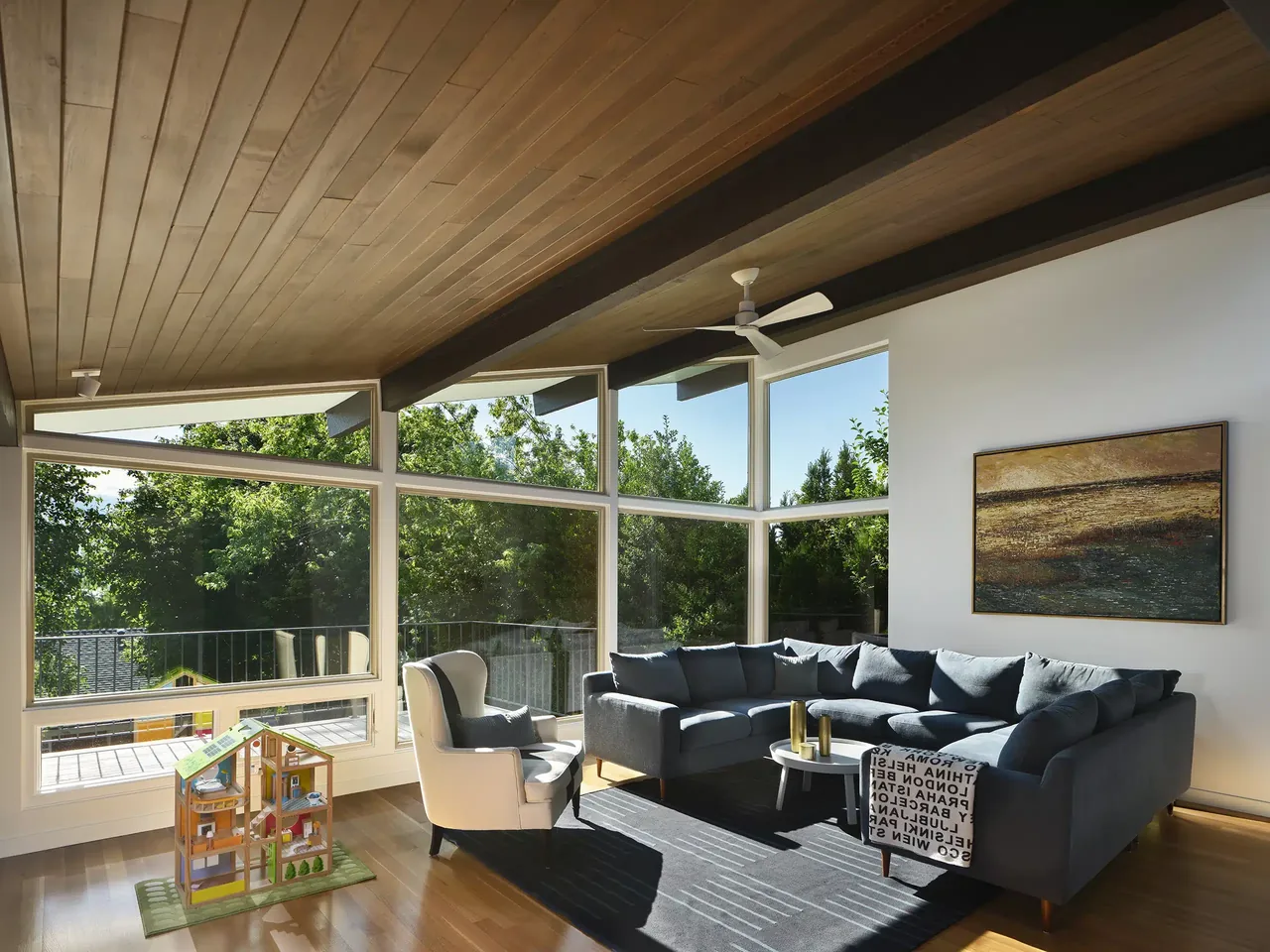 Living room features expansive windows with forest views, a sectional sofa, and wood ceiling details in a mid-century modern home.