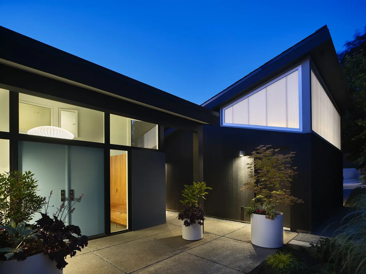 Magnolia Mid-Century Modern home with black wood siding, large illuminated vertical windows, and a sloped roof at dusk.