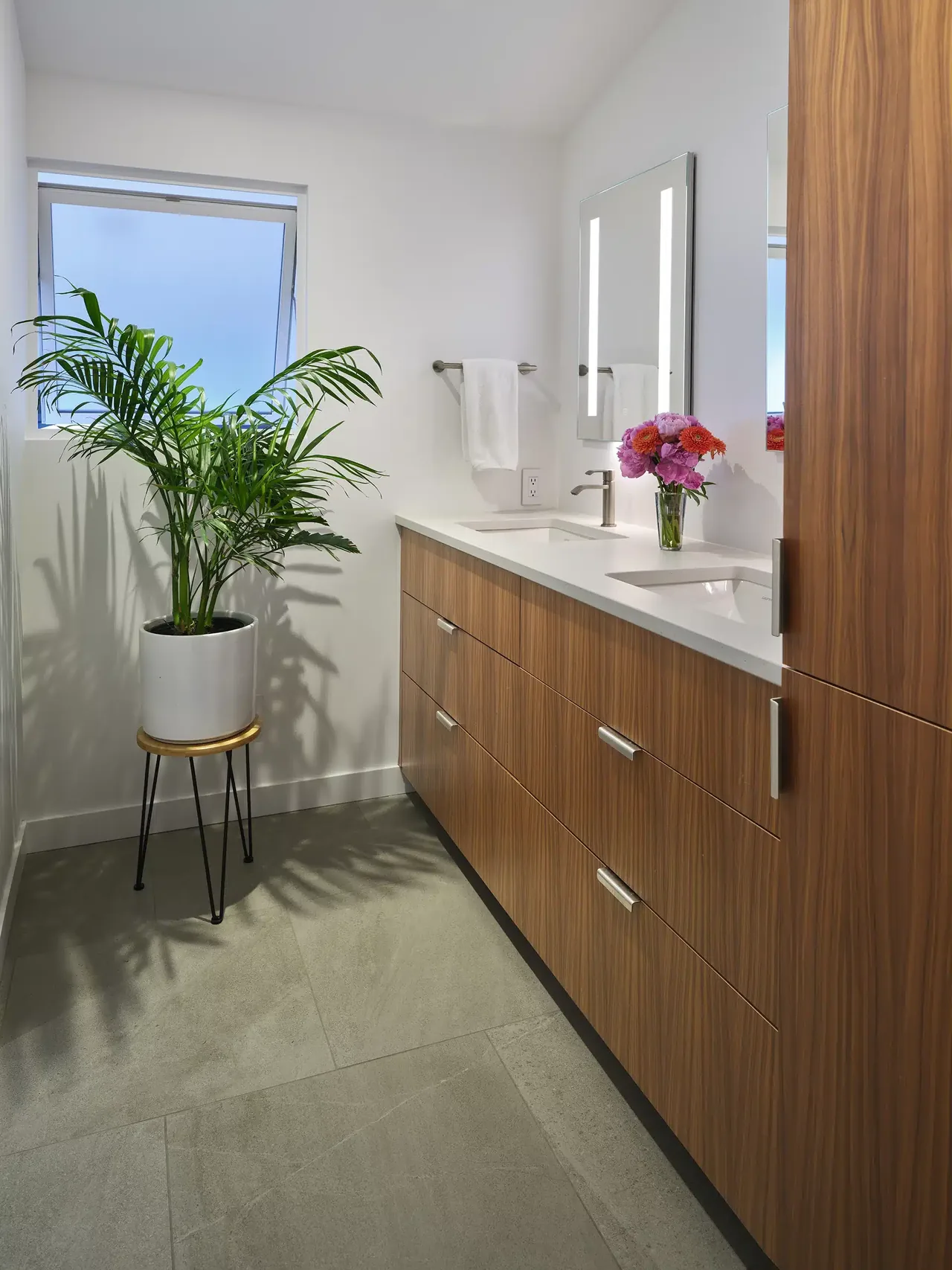 Bathroom featuring a wooden double-sink vanity and backlit mirrors with a potted plant near the window.
