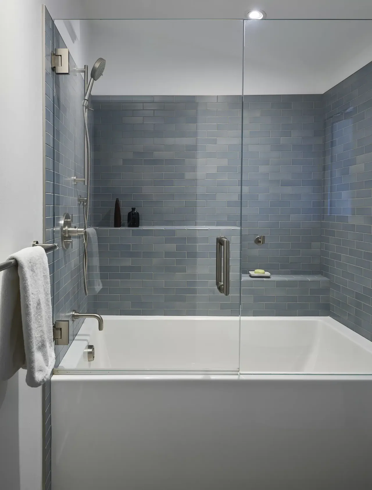 Bathroom shower area with gray subway tiles, a built-in bathtub, a glass partition, and brushed metal fixtures.