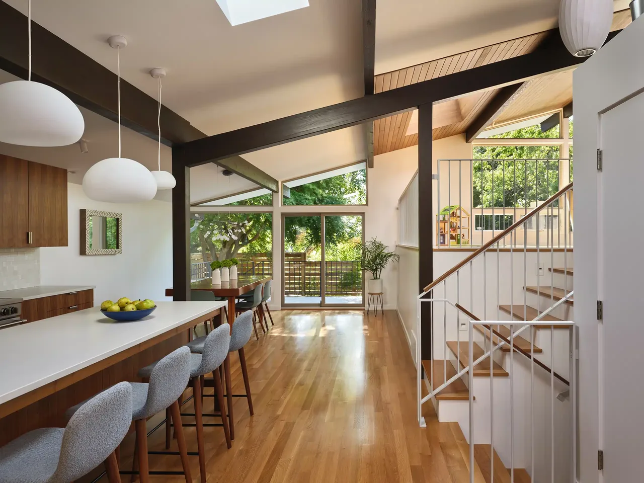 Mid-century modern kitchen and dining space with vaulted ceiling, exposed black beams, and floor-to-ceiling windows opening to outdoor area.