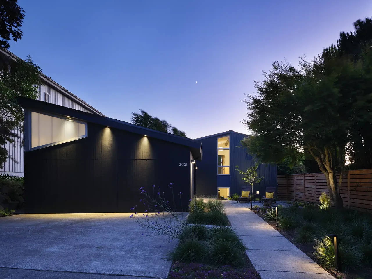 The Magnolia Mid-Century Modern house features angled shed roofs and black exterior walls illuminated by exterior lighting along the pathway.