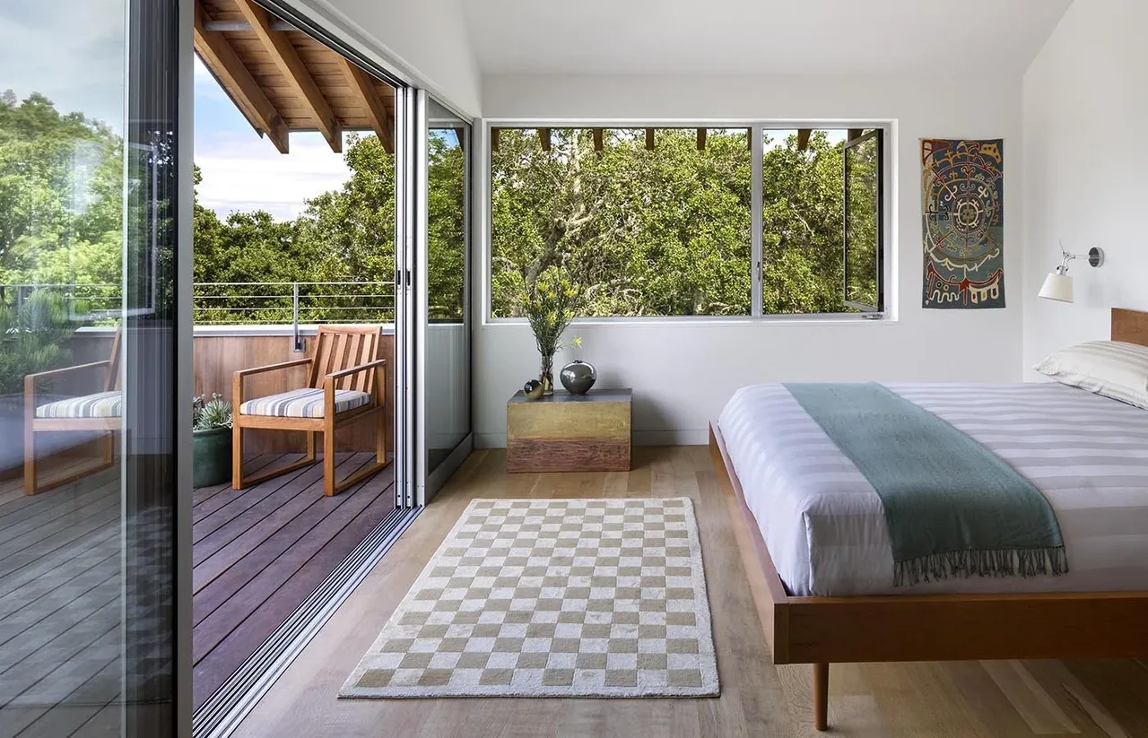 This bedroom features a sliding glass door opening to a balcony with wood furniture and a large window overlooking trees.