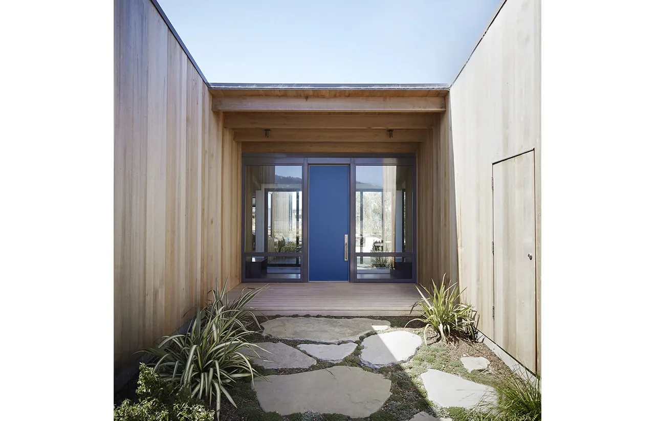 The entrance courtyard features a blue pivot door set within wood siding walls and a stone path with natural plants.
