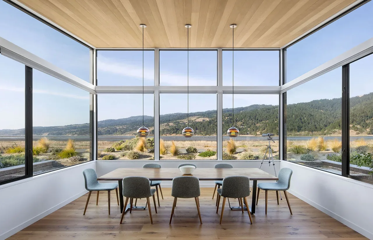 Dining room with wood-paneled ceiling and floor-to-ceiling corner windows overlooking a waterfront view.