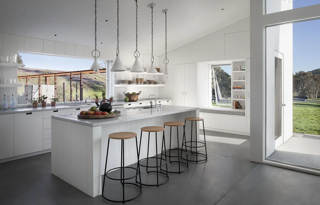 The kitchen in Hupomone Ranch features a large island with seating, white cabinetry, pendant lighting, and expansive windows overlooking the landscape.