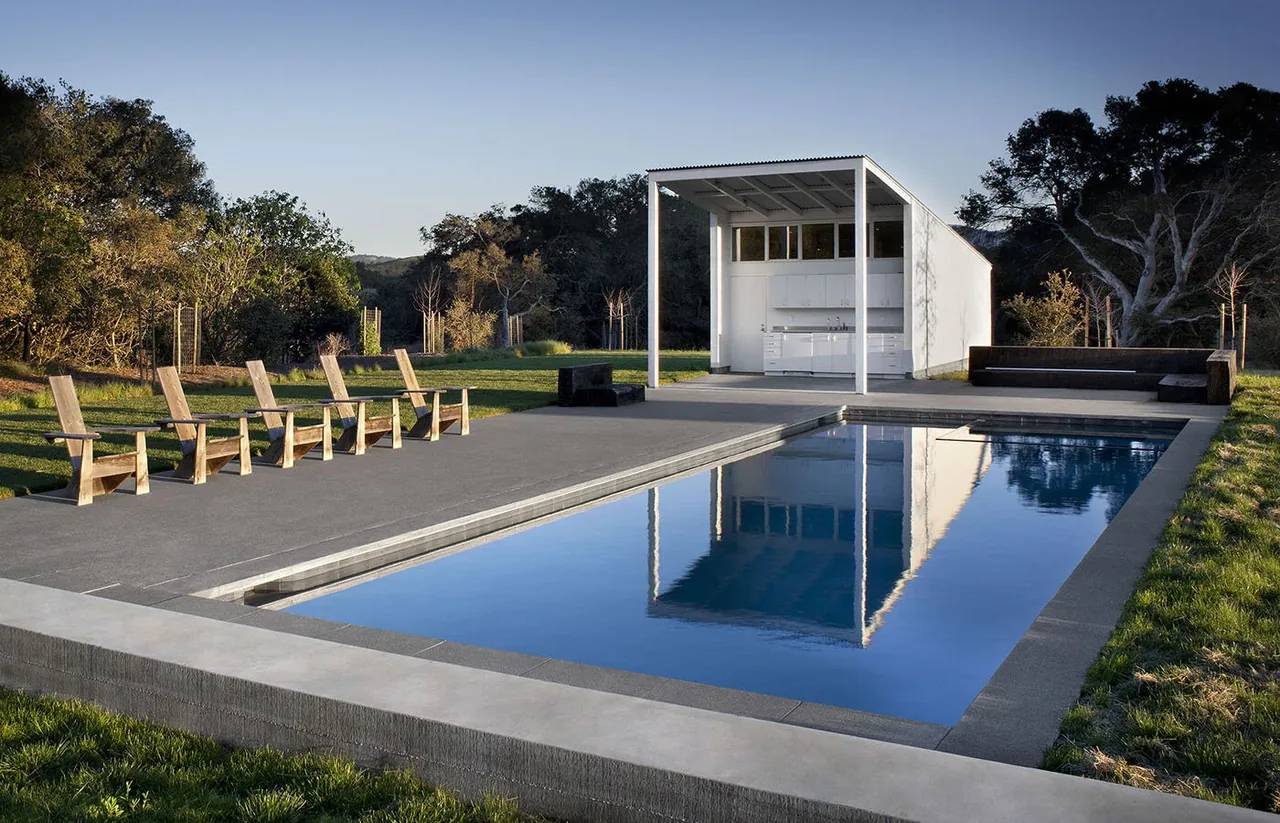 Pool house with open kitchen area next to a rectangular pool on a rural grassy site with mature trees.