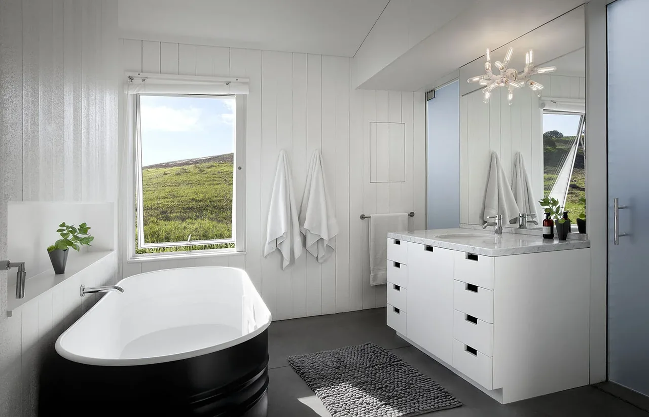 Bathroom features a black freestanding tub, white vanity with marble countertop, and large casement window overlooking a grassy hillside.