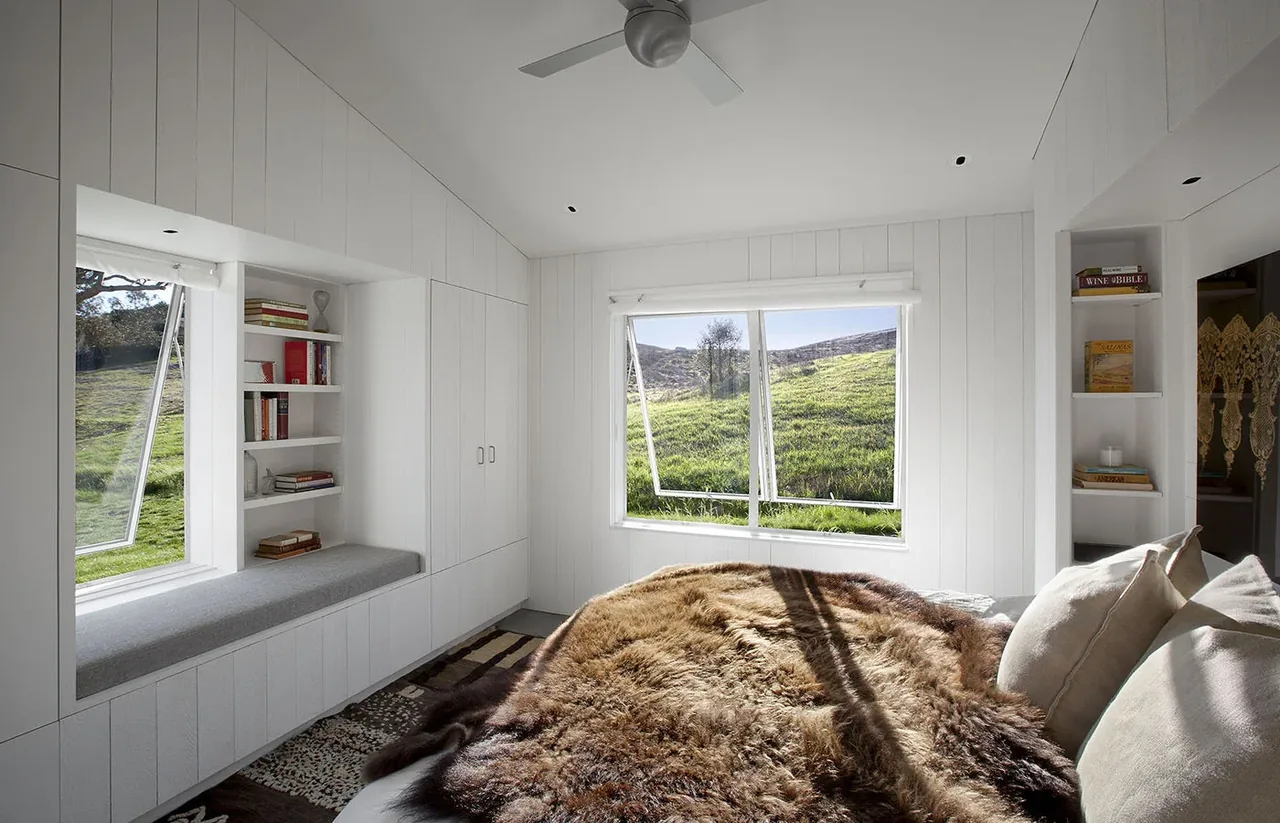 Bright bedroom features white wood paneling, a cozy window seat with shelving, and large casement windows overlooking green hills.