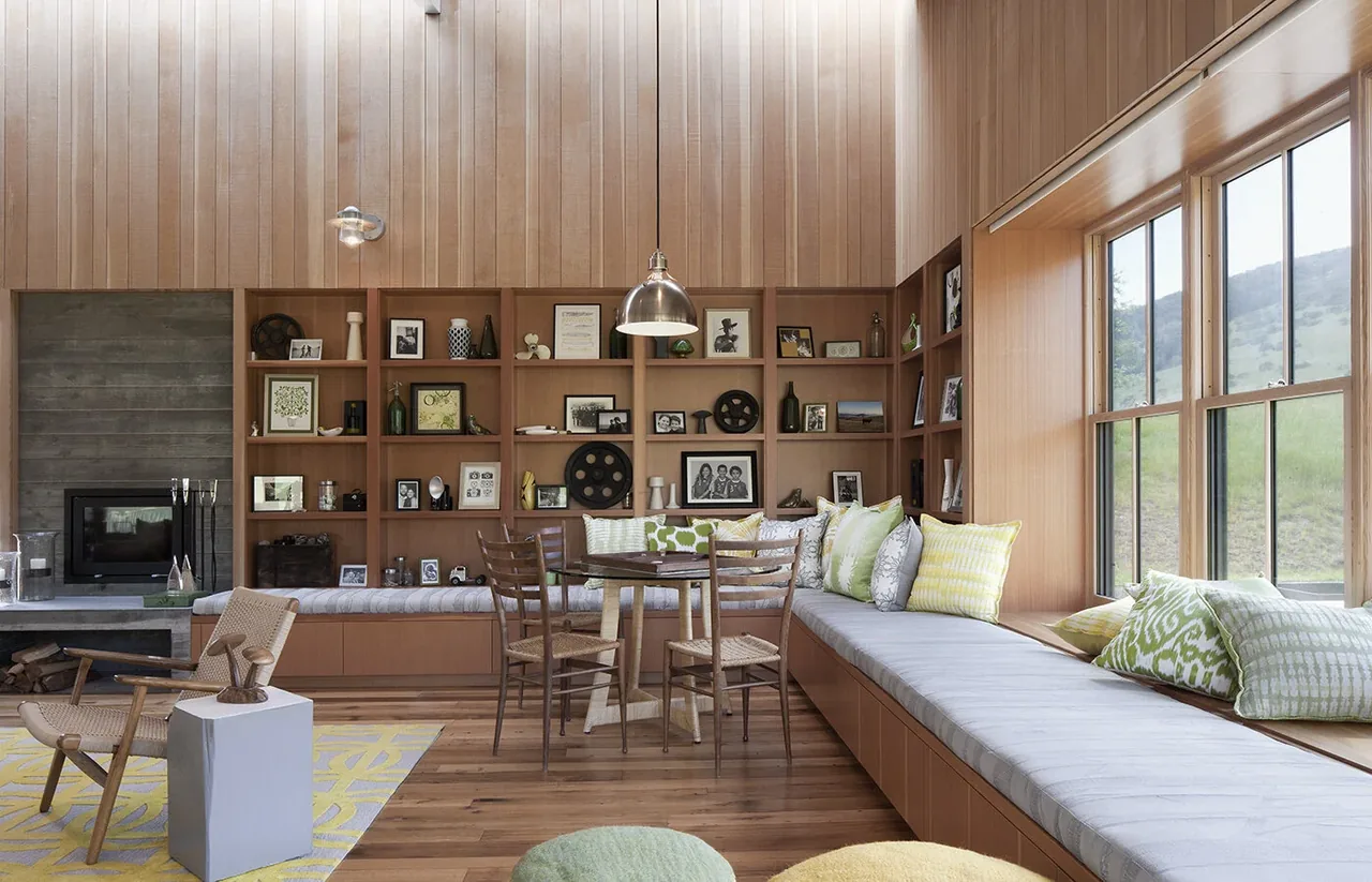 Living room with wood shelves, a window seat, and a fireplace, featuring wood panel walls and large windows overlooking a hillside.