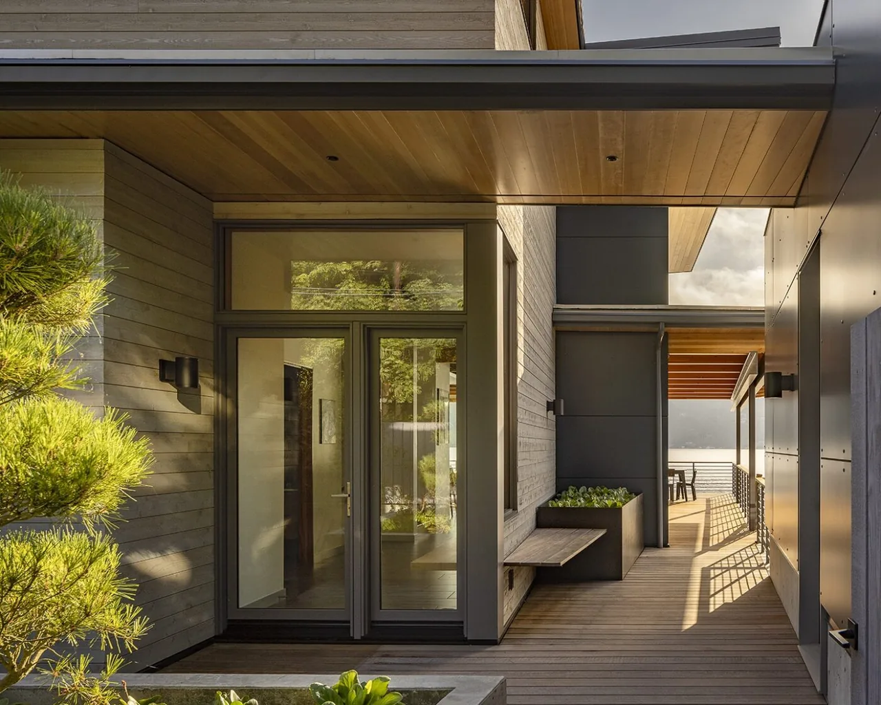 Modern covered deck entrance with wood siding, glass door, and built-in bench overlooking water beyond.