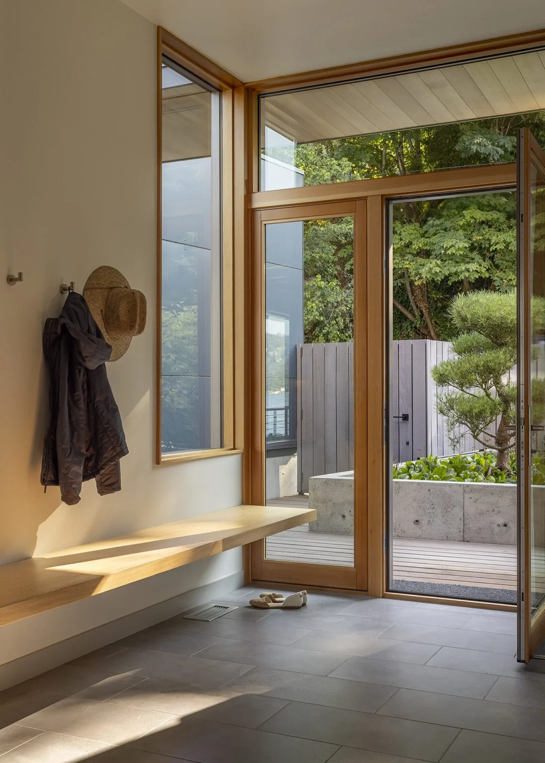 Entry space with a floating wood bench and large wood-framed glass pivot door opening to an outdoor landscaped yard.