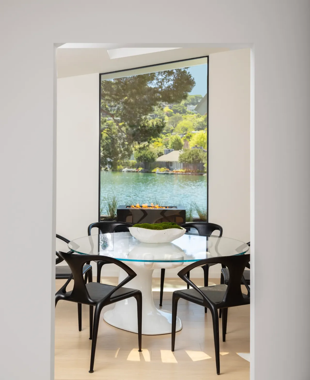 Dining room featuring a glass round table and black chairs with a large window framing a waterfront and outdoor fire feature