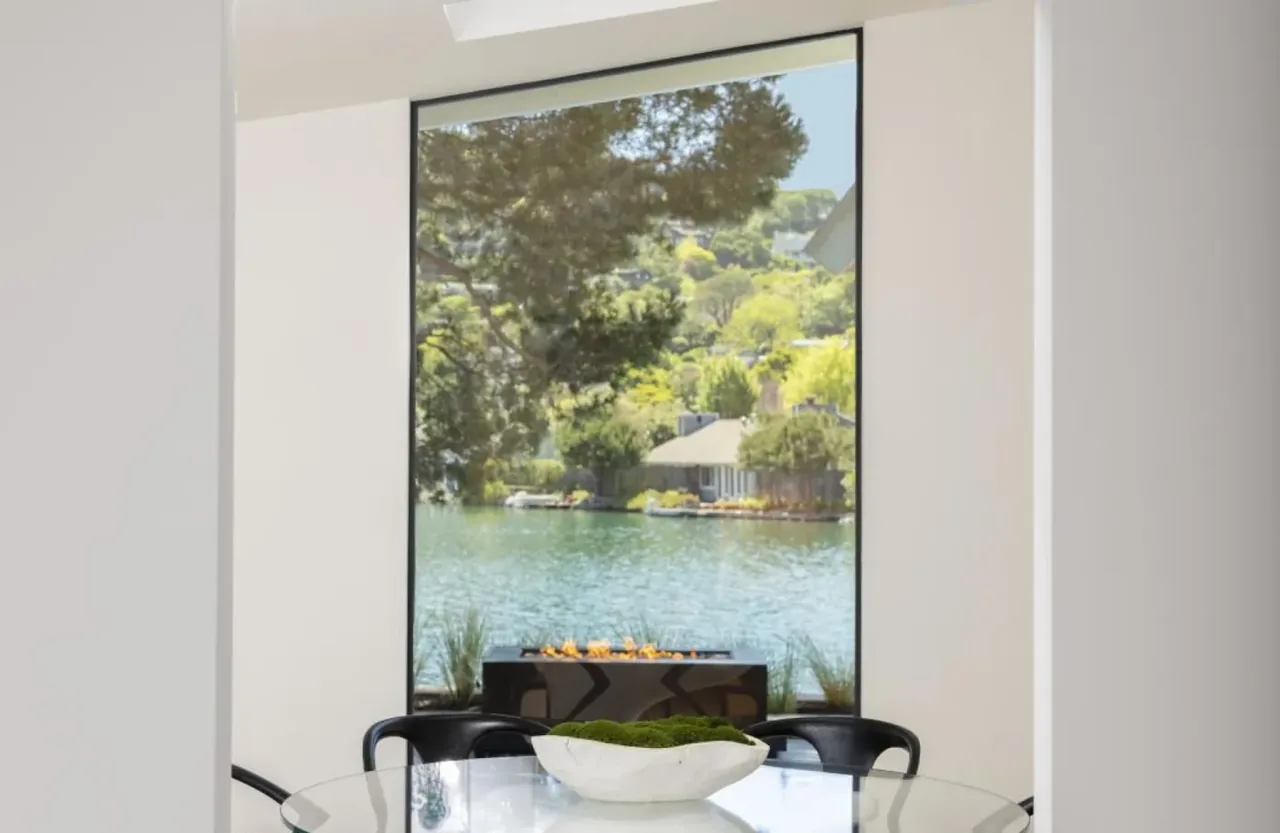 Interior dining area with a large floor-to-ceiling window framing a waterfront scene and modern fireplace.