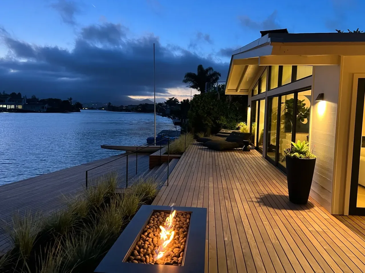 Waterfront wooden deck with recessed fire pit and outdoor seating at dusk, by Orla Studios Architects.