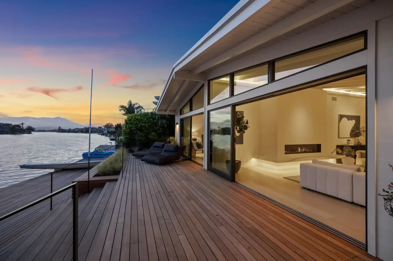 Waterfront wooden deck with sliding glass doors opens to a modern living room featuring a linear fireplace.