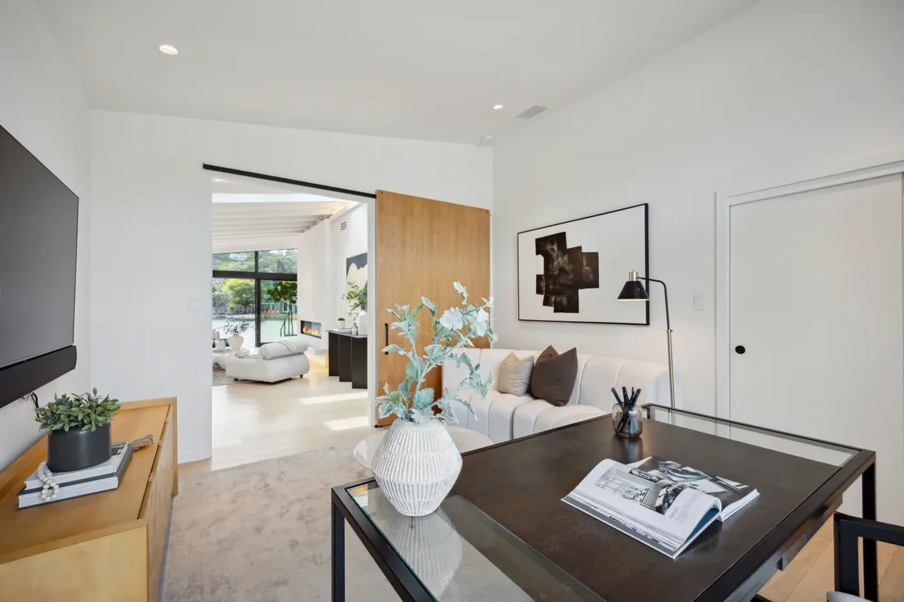 Home office with a glass-top desk and wooden pivot door opens to a living room featuring waterfront views and fireplace.