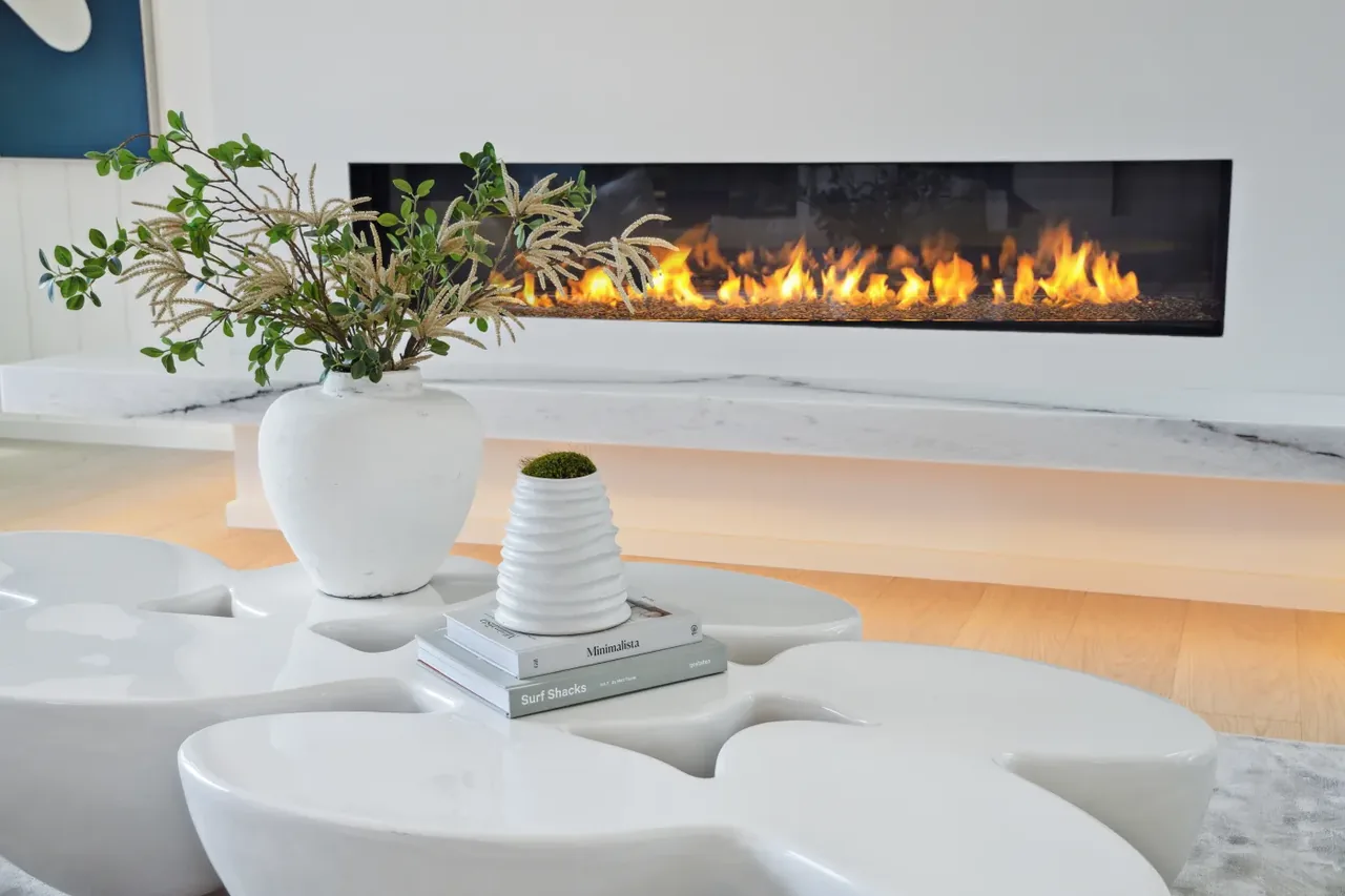 Linear fireplace set within a white wall above a floating marble shelf in a living room with a sculptural coffee table.